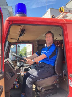 Ein Mann sitzt am Steuer eines roten Feuerwehrfahrzeugs und l&auml;chelt in die Kamera.