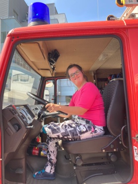 Eine Frau sitzt am Steuer eines roten Feuerwehrfahrzeugs und l&auml;chelt in die Kamera.