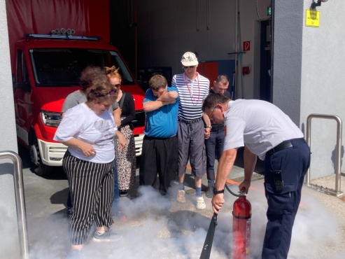 Feuerwehrmann zeigt einer Gruppe von Menschen den Umgang mit einem Feuerl&ouml;scher vor.