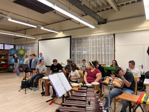Eine sitzende Gruppe von Menschen mit verschiedenen Musikinstrumenten und daneben steht eine Tanzgruppe.