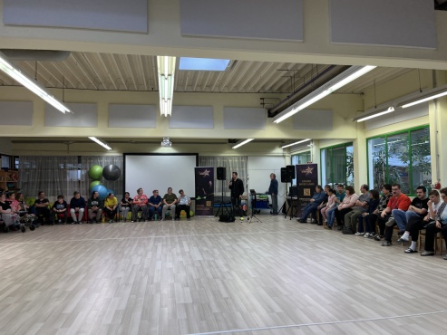 Eine sitzende Gruppe von Menschen hört einem Mann beim Singen zu.