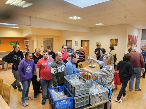 Eine Gruppe von Menschen steht in der Reihe und bekommt Geschenksackerl von einer Frau &uuml;berreicht.