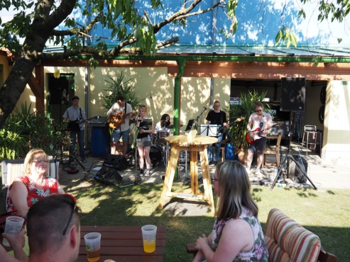 Eine Band musiziert auf verschiedenen Instrumenten im Garten vor einem Publikum.