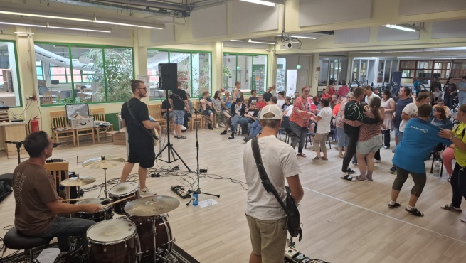 Drei Musiker spielen auf verschiedenen Instrumenten, während einige Personen tanzen.