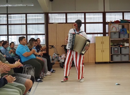 Ein Clown musiziert auf einer Harmonika vor einem sitzenden Publikum.