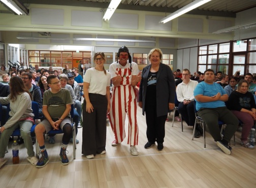 Ein Clown und 2 Frauen stehen vor einem sitzenden Publikum und l&auml;cheln in die Kamera.