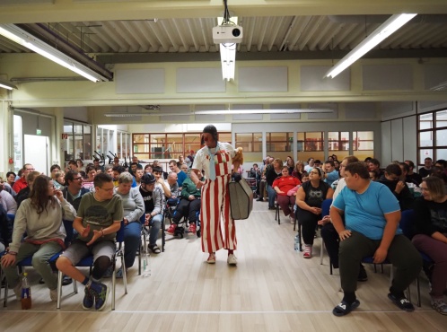 Eine Clown steht in der Mitte zwischen 2 sitzenden Reihen von Menschen.