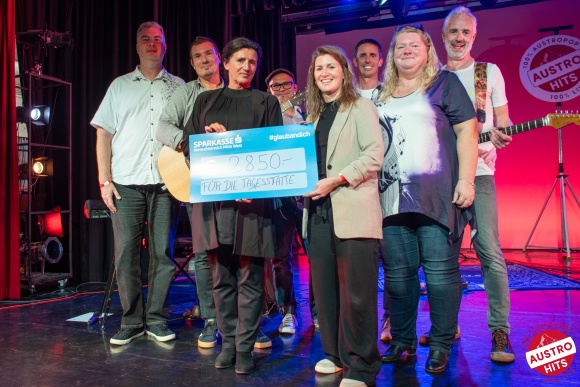 8 Personen auf einer Bühne lächeln in die Kamera. Sie halten einen übergroßen Scheck und 2 Gitarren.