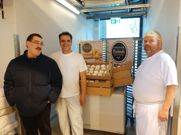 3 Personen stehen vor Schachteln mit Krapfen und lächeln in die Kamera.